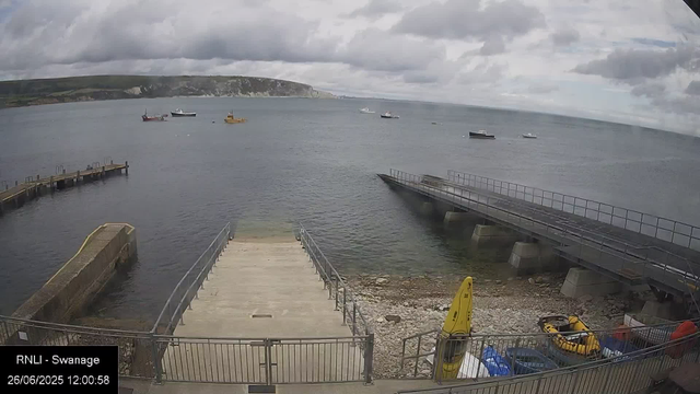 A view of a harbor with several boats anchored on the water and a breakwater extending into the sea. There are cloudy skies overhead, and a distant coastline with cliffs is visible in the background. In the foreground, there is a concrete pier, some safety equipment, and a small inflatable boat. The timestamp indicates the date as June 26, 2025, at 12:00:58.