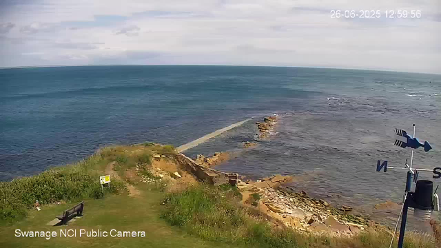 A view of a rocky coastline with a small grassy area and a bench in the foreground. The sea extends to the horizon under a partly cloudy sky. There is a weather vane on the right side of the image, and a sign is visible near the grassy area. The timestamp in the upper right corner indicates it was taken on June 26, 2025, at 12:59:56.