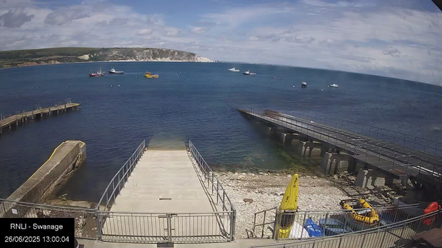 A view of a harbor with a concrete pier extending into the water, surrounded by a metal railing. To the right, a ramp leads down into the water. Several yellow and orange rescue boats are on the rocky beach area. In the background, multiple boats are anchored on the calm, blue sea, with a distant cliff covered in greenery and white chalk cliffs under a partly cloudy sky.