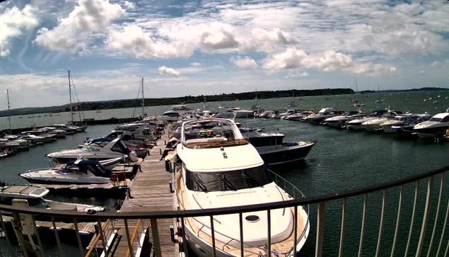 A marina filled with numerous boats and yachts docked along wooden piers. The water is calm, and the sky is partly cloudy with patches of blue. In the foreground, a large white yacht is prominently visible, with a curved railing partially framing the scene.