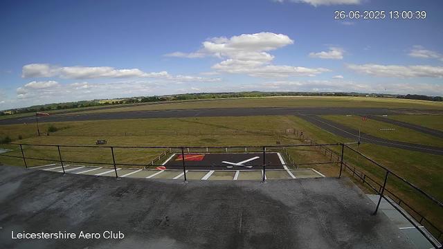 A view from a webcam at Leicestershire Aero Club showing a clear day with a few clouds in the sky. The image captures the runway extending into the distance, surrounded by green fields. In the foreground, there is a helipad with a white helicopter silhouette and a red and yellow marking. The date and time displayed are 26-06-2025 at 13:00:39. The bottom left corner of the image has the text "Leicestershire Aero Club."