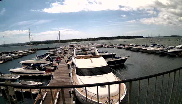 A scenic view of a marina with a wooden dock filled with various boats and yachts. The boats are moored on either side of the dock, and some are covered with tarps. The sky is partly cloudy, with patches of blue visible, and the water is calm. In the background, there is a distant shoreline with trees and landmass extending across the horizon. A metal railing is visible in the foreground, suggesting the photo was taken from a balcony or vantage point overlooking the marina.