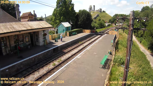 A small train station platform with a building on the left, featuring stone walls and a tiled roof. Two people are standing on the platform. There are green benches and potted plants along the fence. Overhead, there are electrical lines supported by wooden poles. Beyond the platform, there are railway tracks running into the distance. On the hill in the background, on top of a grassy slope, there are ruins of a castle with tall stone walls and towers. The sky is partly cloudy, and the area is surrounded by lush green trees and vegetation.