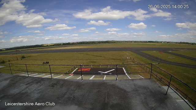 A view from a webcam showing the Leicester Aeroclub airfield under a partly cloudy sky. In the foreground, there is a small platform with a railing, and a marked helipad with a large white "H" and red and yellow markings. The background features a large open grassy area, a runway, and a rural landscape with scattered trees and fields. The timestamp in the upper right corner indicates the date and time as June 26, 2025, at 14:00:25.