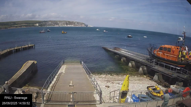A view of a harbor with boats floating in the water, a pier on the left side, and a rescue boat with crew members on the right. There is a concrete slipway leading into the water in the foreground, with a yellow kayak and some gear stored nearby. A person is standing at the railing on the right, looking towards the sea, and another person is on the left walking along the ldeck. The background features cliffs and a cloudy sky.