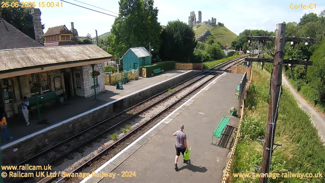 A small train station platform with a few people present. One person in a purple shirt and black shorts is walking away from the camera, carrying a yellow bucket and a green watering can. Another person in dark clothing is standing near a green bench under a shelter made of stone walls and a sloped tiled roof. There is a small teal-colored building, benches, and flower pots along the platform. Overhead power lines run above the railway tracks that run parallel to the platform. In the background, a hillside with ruins or stone structures sits atop a grassy hill, surrounded by trees and greenery. A dirt path runs along the right side of the image, with tall grass and bushes. There are wooden fences and telegraph poles along the platform. The scene is bright and sunny.