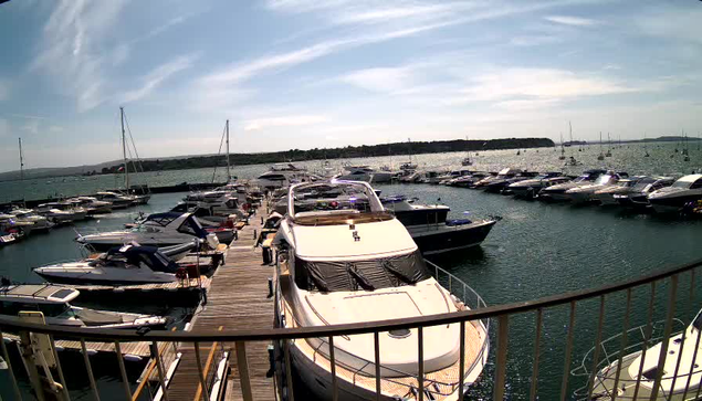 A marina filled with docked boats and yachts, with a wooden pier extending into the water. The sky is partly cloudy, and in the background, there is a hilly shoreline. The boats vary in size and design, and the water is calm, reflecting the sky and boats.