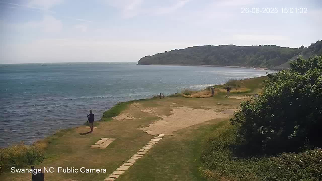 A view of a coastal area with a grassy and sandy shoreline. Several people are walking along the edge of the water, some standing and enjoying the scenery. In the background, there is a large hill or small mountain covered in greenery, extending into the distance. The sky is partly cloudy, with patches of blue visible. A timestamp in the top right corner indicates the date and time as June 26, 2025, at 3:01 PM. The bottom left corner has text that reads "Swanage NCI Public Camera."