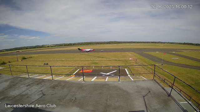 The image shows a view from a webcam at Leicestershire Aero Club. In the foreground, there is a section of the roof with a railing and a designated aircraft parking area marked with a white "X" and red and yellow markings. Beyond that, a small tarmac area with rustic benches is visible. In the background, a small airplane is taxiing on the runway, with two individuals walking nearby. The surrounding landscape features open grass fields and the sky, which is partly cloudy with some areas of blue. The timestamp indicates the date and time as June 26, 2025, at 16:00:12.