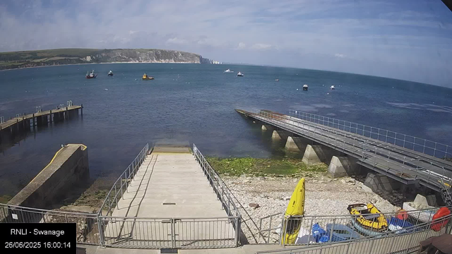 A view of a seaside area with a concrete pier extending into the water, surrounded by fencing. There are some small boats anchored in the water. In the foreground, there are yellow and orange rescue equipment, including a kayak, a small car, and life jackets. The background features a large cliff with grassy slopes and a partly cloudy sky. The timestamp indicates the photo was taken on June 26, 2025, at 16:00.
