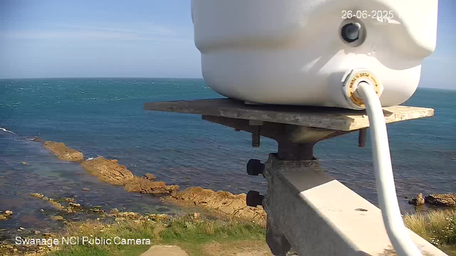 A view of a seaside landscape with rocky shoreline and calm blue ocean under a partly cloudy sky. In the foreground, a white weather instrument or camera is mounted on a wooden platform, with a cable connected to it. The photo is dated 26-06-2025 and has a label "Swanage NCI Public Camera" at the bottom left corner.