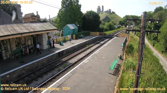 A small train station with a few passengers on the platform. There is a building with a stone facade and a corrugated roof on the left side, where some people are sitting and standing. A woman in a purple shirt is near the entrance, and a man in a white shirt is walking across the platform. A green bench is placed along the edge of the platform, and a little turquoise shed is behind the bench. Overhead, there are power lines, and on the right side, a row of tall wooden electric poles with wires extends into the distance. In the background, a hill with ruins or old structures on top is visible, surrounded by lush green trees and foliage. The timestamp and website information are displayed in the corners of the image.