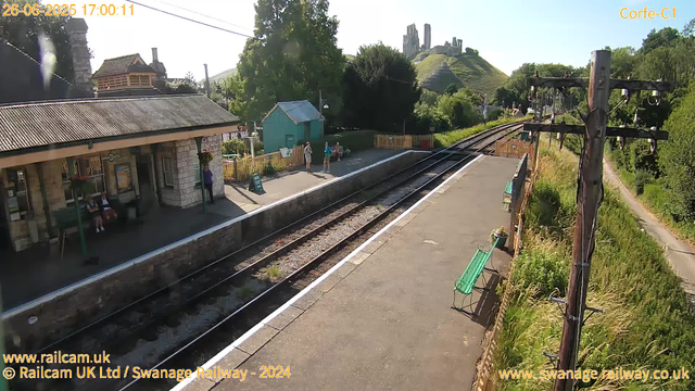 A small train station with a platform and a building on the left side. There are a few people waiting near the platform, some sitting on benches and others standing. The background shows a green hill with some old ruins or castle remains on top. There are trees and a clear blue sky, indicating a bright and sunny day. The railway tracks run through the middle of the image, and there is a tall wooden telephone pole with multiple wires on the right side. The station has some decorative green benches and flower pots along the platform.