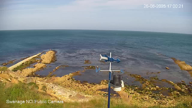 A view of a rocky shoreline extending into the sea under a partly cloudy sky. In the foreground, there is a weather station with an anemometer and other instruments mounted on a pole. The sea appears calm, and a small pier or jetty extends into the water on the left side. The image has a timestamp in the top right corner showing the date and time as June 26, 2025, at 5:00 PM, and a caption in the bottom left corner that reads "Swanage NCI Public Camera."