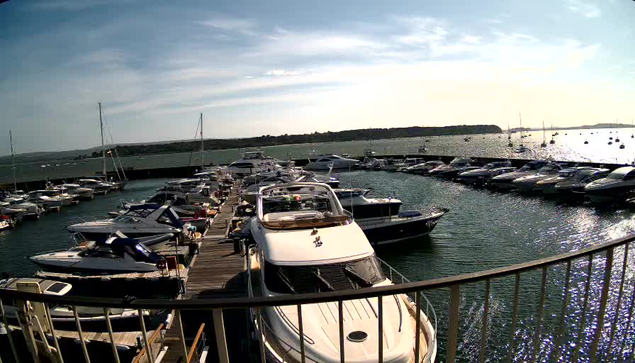 A marina filled with numerous boats and yachts docked along wooden piers, with the calm water reflecting the sky and sunlight, extending towards a distant shoreline under a partly cloudy sky.