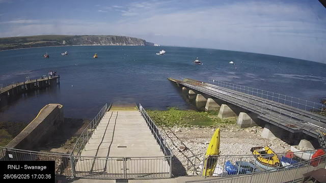 An image of a seaside scene taken from a webcam, showing a clear view of the coast with a pier extending into the water on the left, a ramp leading down to the beach area in the center, and several boats floating on the water in the distance. The sky is partly cloudy, and there is green moss or seaweed near the shoreline. In the foreground, there are parked vehicles and yellow safety equipment. The timestamp indicates the photo was taken on June 26, 2025, at 17:00:36.