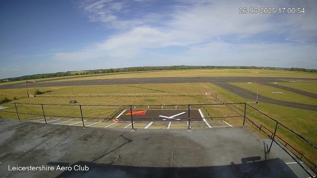 A view from a webcam at Leicesterhire Aero Club showing a clear sky with scattered clouds. The foreground features a small helipad with white painted markings and a red warning zone. Beyond the helipad, there is a grassy area enclosed by a wooden fence, and a small picnic table is visible. In the distance, there is a runway with a red vehicle parked along its side, and the surrounding landscape is open fields with some trees on the horizon. The timestamp in the upper right corner reads "26-06-2025 17:00:54".