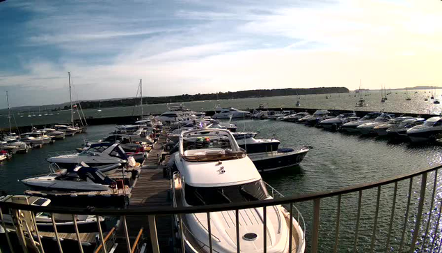 A marina filled with various boats and yachts docked along wooden piers, with a calm body of water extending into the distance under a partly cloudy sky.
