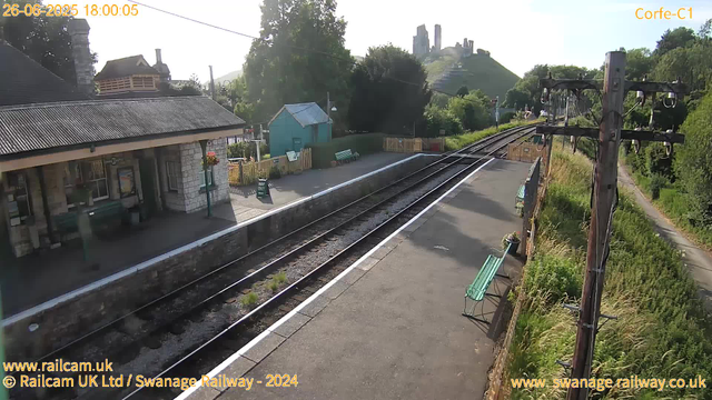 A small train station platform with a stone building on the left, a green bench and a small blue shed in the background. Railway tracks run through the scene, with railway overhead wires and support poles on the right. In the distance, a hill with ruins or old structures is visible, and the scene is bathed in sunlight, suggesting late afternoon. There are flowers and greenery along the right side of the platform. The date and time are displayed at the top left corner.