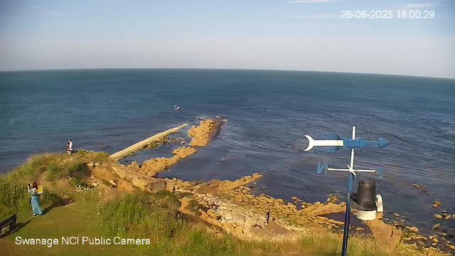 A coastal scene showing a rocky shoreline with a small pier extending into the ocean. There are three people near the water, two on the grassy area and one on the rocks. An small boat is visible in the distance on the water. The sky is clear with no clouds, and the date and time in the corner indicate it is June 26, 2025, at 6:00 PM. A weather vane with directional markers (N, S, E, W) is prominently positioned in the foreground. The image is from a webcam labeled "Swanage NCI Public Camera."