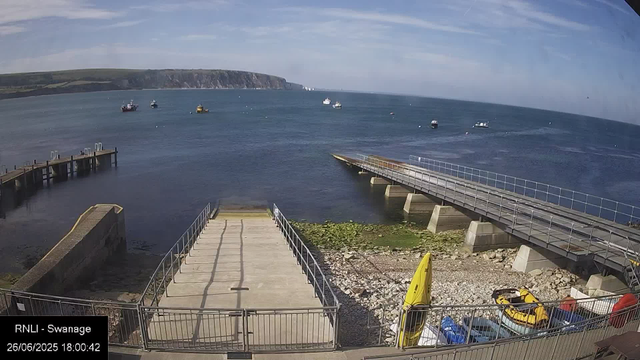 A coastal scene viewed from a webcam showing a pier leading into the sea, with several boats floating on the water. In the background, there are cliffs under a mostly clear sky with some scattered clouds. In the foreground, there are some rocks, a yellow lifeguard buoy, and parked cars. The image is timestamped "26/06/2025 18:00:42" and labeled "RNLI - Swanage".