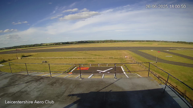 A view from a webcam at Leicesterhire Aero Club shows a clear day with a bright blue sky and some scattered clouds. In the foreground, there is a small helicopter landing pad with a white helicopter silhouette painted on it, surrounded by safety barriers. Beyond the landing area, there is an expansive grassy field and a paved runway stretching into the distance. The timestamp on the image reads 26-06-2025 18:00:50.
