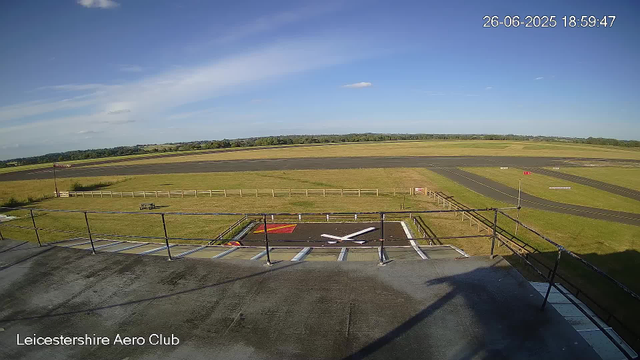 A clear view of an empty airplane parking area at the Leicesterhire Aero Club with a runway in the background. The sky is mostly sunny with some light clouds, and the time and date displayed in the upper right corner is June 26, 2025, 18:59:47.