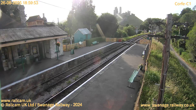 A small railway station platform with a stone building on the left side, featuring a corrugated roof and a hanging flower basket. There are two green benches and a sign that reads "Way Out." The train tracks run through the center, curving gently to the right in the distance. On the right side, there are old wooden railway poles with wires and some greenery and trees alongside a narrow path. In the background, a hill with a castle or large structure on top is visible above the trees. The sky is bright and sunny, with sunlight casting a slight glare. The image timestamp shows the date as 26-06-2025 at 18:59:53.