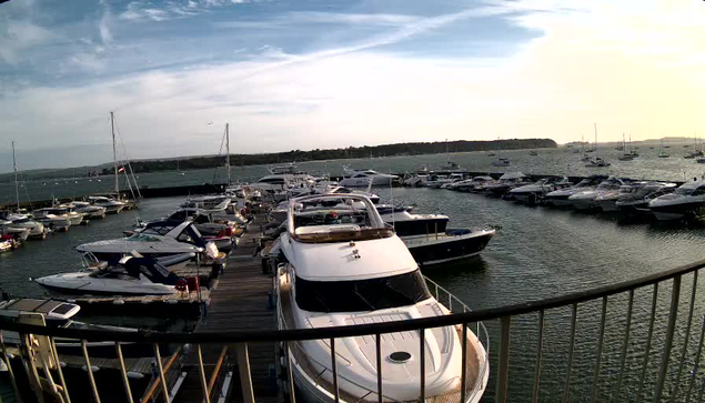 A marina filled with various boats and yachts docked along wooden piers, with calm water reflecting the vessels. The sky is partly cloudy with patches of sunlight. A curved railing is visible in the foreground.