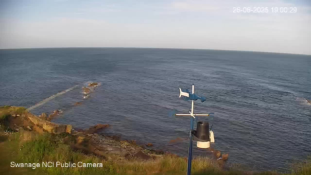 A view of the ocean coastline during daytime, with gentle waves and a few rocks near the shore. In the foreground, there is a small weather vane mounted on a pole, and some greenery along the edge of the land. The sky is clear with minimal clouds. The timestamp in the upper right corner indicates the date and time as June 26, 2025, at 19:00:29. The bottom left corner has a label reading "Swanage NCI Public Camera."