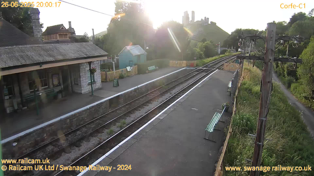 A quiet railway station platform at sunset with three benches, a small blue building and a stone station building. A railway track runs through the scene, with wires and a wooden pole on the right. In the background, green hills and ruins of a castle are visible. The sky is bright as the sun sets behind the trees and hills, creating a warm glow and lens flare effect. The image is timestamped "26-06-2025 20:00:17".