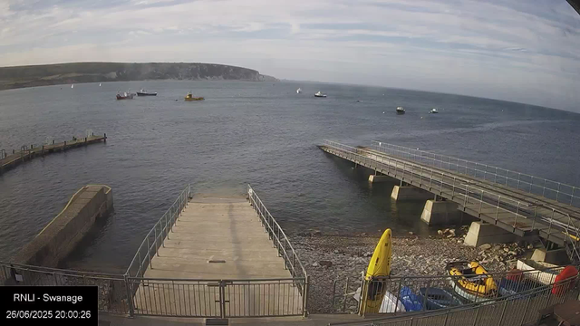 A view of the harbor at Swanage, with a concrete pier extending into the water in the foreground. Several boats are anchored in the bay, with a grassy coastline and cliffs in the distance. The sky is partly cloudy.