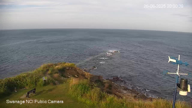 A view of a coastline taken from a webcam. In the foreground, there is a grassy area with a bench and a person standing near it. The rocky shoreline extends into the ocean, which has gentle waves. A weather station with anemometer and wind vane is visible on the right side of the image. The sky is overcast with light clouds. The date and time displayed in the top right corner are "26-06-2025 20:00:39".