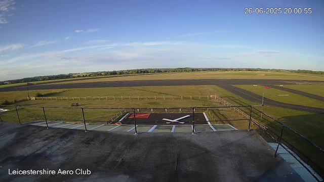 An outdoor scene showing the Leicester Aero Club with an airstrip in the distance. The sky is clear with a few clouds, and the time and date displayed in the top right corner are June 26, 2025, at 20:00:55. In the foreground, there is a rooftop with a large airplane propeller logo painted on it, surrounded by a black railing. The surrounding area is grassy with some wooden fencing and minimal equipment, and the landscape extends to distant fields and trees.