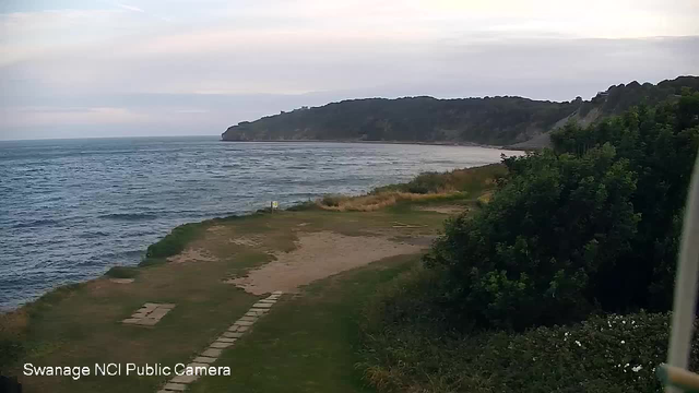 A view of a coastal landscape with a grassy area, a narrow paved pathway leading toward the beach, and a sandy shoreline. The ocean has gentle waves, and there is a large, green, tree-covered headland in the background under a partly cloudy sky.