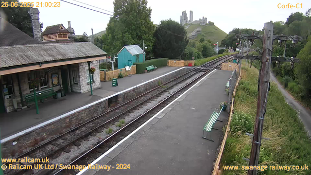 A small train station platform with two green benches, a blue small building, and some potted plants. There are wires and poles along the platform, and a railway track running parallel. In the background, there is a grassy hill with the ruins of a castle on top. The sky is overcast, and the timestamp indicates it is evening. The scene appears peaceful and rural.