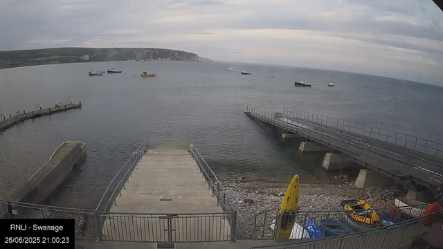 A view of a harbor with a cloudy sky. There are several boats floating on the water, including a sailboat and a yellow boat. In the foreground, there are concrete piers and a small concrete dock extending into the water, with a yellow kayak and some other equipment stored near the metal railing. The coastline with cliffs is visible in the distance across the bay.