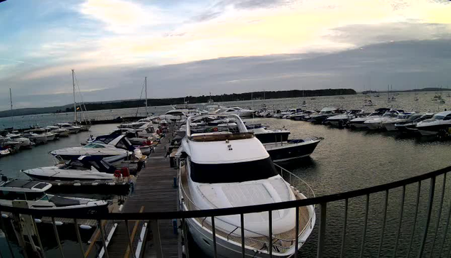 A marina filled with numerous boats and yachts docked along a wooden pier. The sky is partly cloudy with a hint of sunset, and the water appears calm. A black railing is in the foreground.