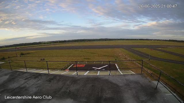 An outdoor view from a webcam at Leicestershire Aero Club showing a grassy airfield with a small helicopter landing pad in the foreground, surrounded by a safety fence. The sky is partly cloudy, and there is a runway in the background with green fields and trees stretching to the horizon. The timestamp in the top right corner reads "26-06-2025 21:00:47".
