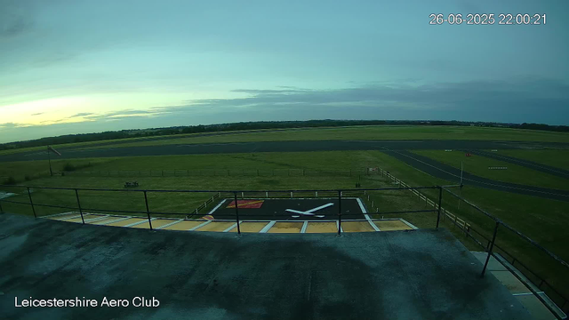 The image shows a view from a webcam at Leicesterhire Aero Club, overlooking an airfield. In the foreground, there is a helipad with a white helicopter symbol painted on it, enclosed by a safety railing. The airfield has a large grassy area with a small flagpole and a windsock. In the background, there are taxiways and runways stretching across the landscape. The sky is mostly cloudy with a hint of sunlight near the horizon, and the timestamp indicates the photo was taken on June 26, 2025, at 22:00:21.