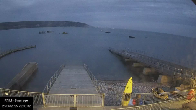 A view of a harbor at dusk, with several small boats anchored in the water. There are two piers extending into the water, and a larger concrete and metal platform in the foreground surrounded by a metal railing. The sea is calm, and a distant shoreline with hills is visible in the background. The timestamp indicates the photo was taken on June 26, 2025, at 10:00 PM.