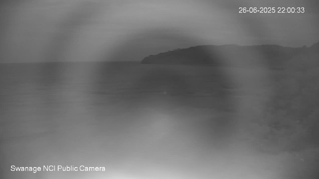 A black and white image from a webcam shows a coastal scene with a large body of water in the foreground and a landmass or island in the distance towards the right. The sky above is overcast with thick clouds. The timestamp in the top right corner reads "26-06-2025 22:00:33," and the bottom left corner notes it as "Swanage NCI Public Camera."