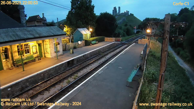 A small railway station platform at dusk, illuminated by warm yellow lights. There are green benches under hanging flower baskets, a small stone building with a green door and window frames, and a sign indicating "Way Out." The train tracks run through the center, with a wooden fence and a narrow canal along one side, and a grassy hillside with ruins atop a hill in the background. An electricity pole with wires is visible near the tracks, and the sky is gradually darkening. The image is marked with the date and time "26-06-2025 22:00:53" and the website information "www.railcam.uk" and "www.swanage railway.co.uk."