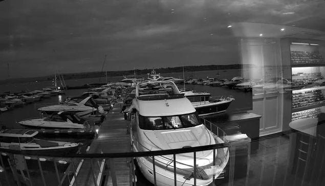 A view from inside a building looking out through a glass window at a marina. Several boats and yachts are docked along a wooden pier, with the water and horizon in the background. The sky appears overcast, and the scene is in black and white. Inside the building, there are shelves or display cases with wine bottles, and a small table is partially visible on the right.