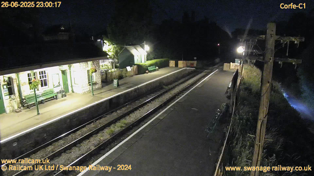 A small train station platform at night, illuminated by bright lights. The platform has green benches, flower baskets hanging from green posts, and a small building on the left with windows lit from inside. There is a "Way Out" sign near the building. Next to the platform are railway tracks, and on the right side, there is a wooden fence with a few objects leaning against a utility pole. The background is dark with some trees and distant structures faintly visible. The timestamp shows the date and time as "26-06-2025 23:00:17", and there are website addresses in the bottom corners.