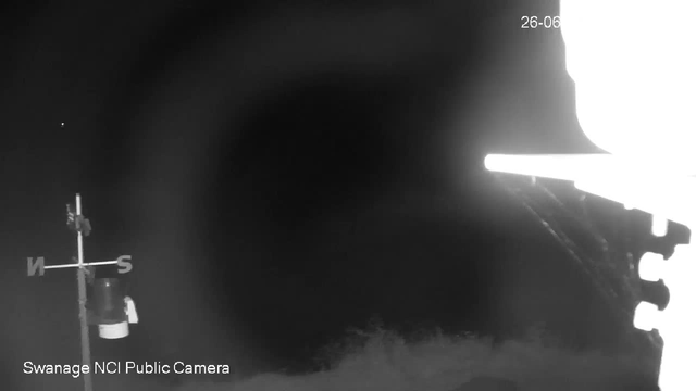 A black and white image from a webcam shows a dark, empty corridor or tunnel with bright light coming from the right side. A weather vane with an arrow and the cardinal directions (N, S, E, W) is visible on the left, along with a small object hanging beneath it. The words "Swanage NCI Public Camera" are written in the bottom left corner. The date "26-06" appears in the top right corner.