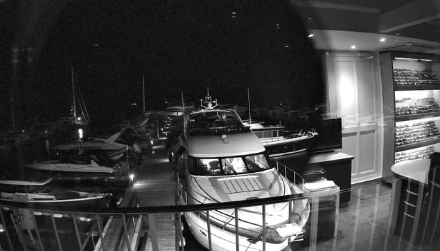 A black-and-white photo taken from a webcam shows a marina at night. In the foreground, there is a large luxury yacht docked closely, with its sleek, curved design visible. To the left, there are several other boats of various sizes, including sailboats and motorboats, moored in the water. The dark night sky above contrasts with the lit-up interior of a room on the right side of the image, which features shelves filled with shelves of bottles and a table with chairs. The scene combines maritime and indoor elements under low light conditions.