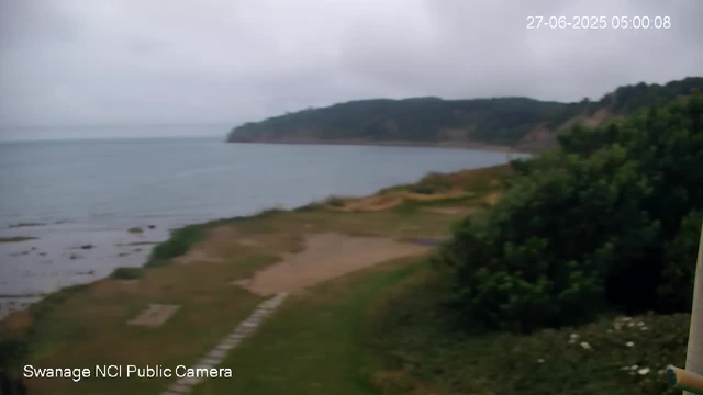 A view of a coastal landscape taken from a webcam. The image shows a shoreline with a grassy and sandy area leading to the water. In the distance, there is a hill or headland covered with greenery. The sky is overcast, giving the scene a subdued and cloudy appearance. The timestamp in the top right reads "27-06-2025 05:00:08," and the bottom left indicates it is from "Swanage NCI Public Camera."