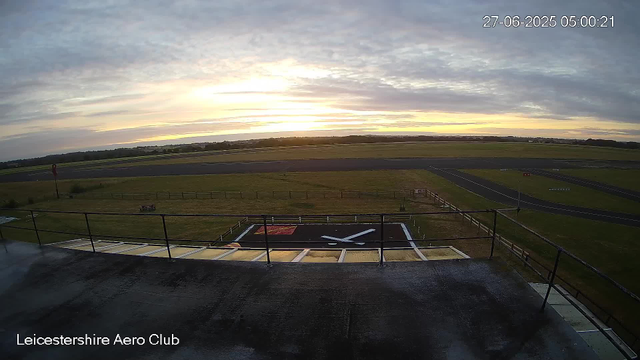 A view from a webcam at Leicestershire Aero Club shows a runway and grassy field under a cloudy sky during sunrise or sunset. In the foreground, there is a helipad with a large white "H" and a small red symbol, and a black railing surrounds the helipad area. The horizon features open fields and scattered trees, with the sun partially obscured by clouds, casting a soft glow across the scene. The timestamp on the image reads "27-06-2025 05:00:21."