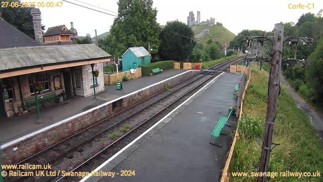 A small railway station platform with a stone station building on the left, featuring hanging flower baskets and green benches. There are three electric poles along the platform, with wires overhead. A paved footpath runs alongside the platform on the right, bordered by a wooden fence and overgrown grass with wildflowers. In the background, a green hill with ruins of an old castle or fortress on top can be seen, surrounded by trees and lush greenery. The sky appears to be overcast or early morning. The image also includes a timestamp and website information at the top and bottom.